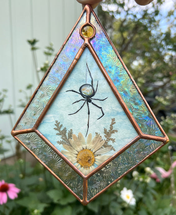 Spider with miniature landscape in body above pressed daisy flower and fern painted with gouache on paper set between glass. Iridescent floral textured glass border and yellow gem.