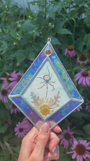 Video of refraction. Spider with miniature landscape in body above pressed daisy flower and fern painted with gouache on paper set between glass. Iridescent floral textured glass border and yellow gem.